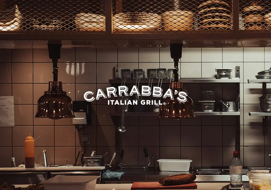 http://Carrabba’s%20Italian%20Grill%20logo%20displayed%20over%20a%20warm,%20dimly%20lit%20kitchen%20scene%20with%20copper%20heat%20lamps%20and%20stainless-steel%20cookware.
